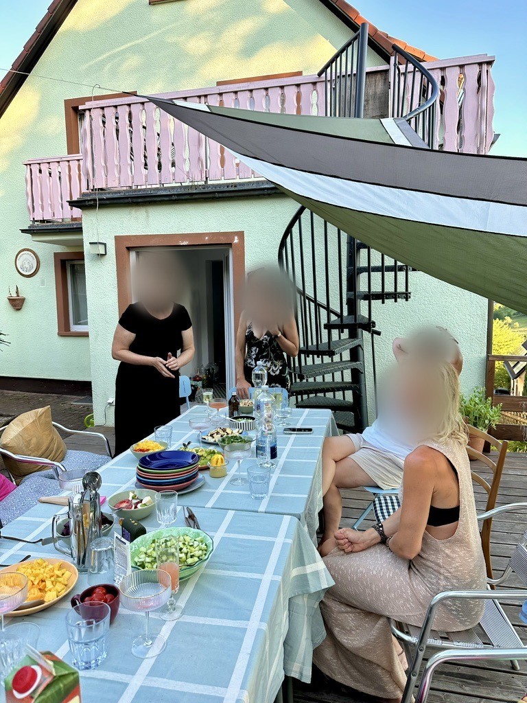 Lange Tafel mit hellblauer Tischdecke und bunten Schüsseln auf der Holzterrasse, daneben die Wendeltreppe